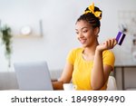 Easy Payment And Cashback Concept. Portrait of excited african american young lady holding credit card in hand and using laptop, sitting at table at home, typing on keyboard, blurred background