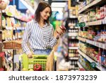Small photo of Portrait Of Smiling Woman With Shopping Cart In Supermarket Buying Groceries Food Walking Along The Aisle And Shelves In Grocery Store, Holding Glass Jar Of Sauce, Choosing Healthy Products In Mall
