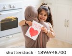 Smiling little girl holding greeting card for Happy Mother's Day with drawn red heart and hugging her muslim mom. Loving islamic family bonding together at home, closeup shot with free space