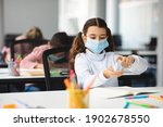 Small photo of Preventive Measures. Small girl applying antibacterial sanitizer spray on hands, wearing disposable protective medical mask, sitting in classroom at school. Stop the spread of infection and germs