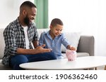 Small photo of Personal Savings. African Father And Son Putting Coin In Piggybank Sitting On Sofa At Home, Dad Teaching His Kid Financial Literacy, Talking About Money And Budget Planning. Selective Focus