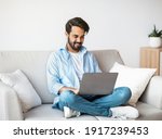 Distant Work. Happy Arab Freelancer Man Using Laptop Computer At Home, Typing On Keyboard, Young Eastern Guy Sitting On Sofa In Living Room, Working Remotely On Online Project, Copy Space