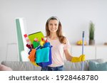 Cheerful millennial housewife holding bucket full of cleaning supplies at modern apartment. Portrait of cheerful young lady with sanitary equipment looking at camera and smiling