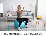 Strong mature woman working out with dumbbells on fitness ball at home, blank space. Fit senior lady taking care of her health, having domestic training, leading active lifesyle, indoors