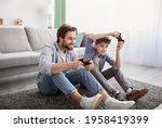 Small photo of Videogames concept. Joyful father and son competing in online games, holding joysticks while sitting on carpet in living room and spending time together on weekend, copy space