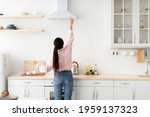 Rear back view of young millennial housewife choosing and selecting mode on range exhaust hood, pushing button on mechanical fan above the stove, standing in modern kitchen at home