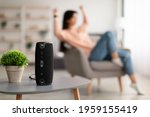 Modern Technology Concept. Closeup of smart portable wireless speaker on the table, selective focus. Young woman listening to music and dancing, sitting on armchair in the blurred background