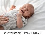 Closeup Portrait Of Crying Little Newborn Baby In Bodysuit Lying On Bed At Home, Upset Infant Child Suffering Colics Or Gas Problems, Loving Mother Making Tummy Massage To Small Kid With Two Hands