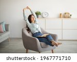 Gorgeous Indian woman stretching in comfy armchair while reading indoors. Attractive young lady with open book, enjoying lazy morning, having peaceful weekend. Stay home hobbies