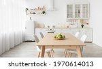 Contemporary minimalist interior of kitchen and dining room. White furniture with utensils and dinner table with chairs in Scandinavian style. Modern light design
