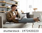 Mobile Communication. Portrait of smiling young asian guy using laptop and talking on mobile phone, holding cup and drinking coffee, working remotely sitting on the couch at home office