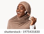 Natural Makeup And Beauty Concept. Portrait of cheerful muslim black lady in hijab holding brush and applying powder on cheek, looking and posing at camera, isolated on white studio background