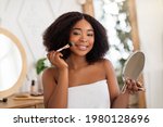 Smiling black woman applying cosmetic powder or blusher on her face, using decorative makeup, looking in mirror at home. Lovely young lady wearing bath towel, putting on cosmetics in morning