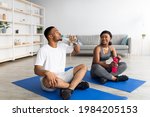 Athletic black couple in sportswear sitting on yoga mats, drinking water from bottles after domestic training, indoors. Cool guy and his girlfriend taking break after workout, staying hydrated
