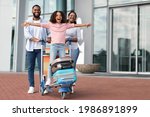 Family Trip Concept. Portrait of cheerful African American girl having fun and spreading hands, ready for vacation, standing on luggage cart. Parents walking with baggage trolley, riding daughter