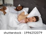 Small photo of Angry young woman shouting and covering ears with pillow while her husband snoring on bed. Millennial lady being disturbed by her boyfriend's snort problem, cannot fall asleep