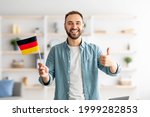 Happy Caucasian man showing thumb up and flag of Germany, posing and smiling at camera indoors. Cheerful millennial student recommending foreign education, learning German language
