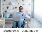 Portrait of happy handicapped black guy with laptop showing thumb up gesture, smiling at camera indoors. Paraplegic young man in wheelchair recommending online job during covid pandemic
