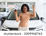 Happy Owner. Overjoyed Black Lady Holding Keys Of Her New Car After Buying, Cheerful African American Woman Standing Near Vehicle In Dealership Center And Raising Fist With Excitement, Copy Space