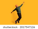 Small photo of Cheerful young african american man in orange hat standing on tiptoes over yellow background, full length shot of happy millennial black hipster guy dancing and having fun in studio, copy space