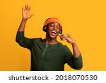 Technologies For Fun. Cheerful black hipster guy listening music in wireless headphones and singing, positive young african man using smartphone as microphone, having fun on yellow background