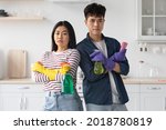Small photo of Serious asian man and woman with cleaning sprays and sponges standing back to bact with arms crossed on chest at white kitchen, looking at camera, posing while house-keeping at home together