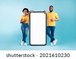 Great mobile app. African American couple pointing at giant smartphone with mockup, promoting application or website, advertising product or service, blue background. Full length