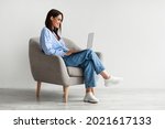 Smiling young woman in casual wear working online, sitting in armchair and using laptop against white studio wall, copy space. Cheerful Caucasian lady surfing internet on portable pc
