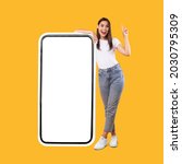 Small photo of Wow. Happy Woman Leaning On Big Smartphone With Blank White Screen And Pointing Finger Up, Cheerful Lady Recommending New App Or Website, Standing On Yellow Background, Mock Up Image, Full Body Length