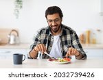 Positive arab guy hipster having healthy breakfast at home, eating fresh vegetables, sausages, drinking coffee, sitting at kitchen table, copy space. Handsome indian man starting day with nice meal