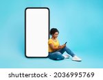 Cheery African American lady sitting near big cellphone with empty white screen, using mobile device, checking new cool app on blue studio background, mockup for website or application design