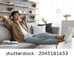 Small photo of Rest And Relax Concept. Calm asian man sitting on couch, listening to music, audio book, podcast, enjoying meditation for sleep and peaceful mind in wireless headphones, leaning back, copy space