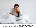 Small photo of Hungry young female greedily eating burger on bed. Eating disorder, consolation with overeating and gluttony, obesity after mental depression, loss substitution with unhealthy food consumption problem