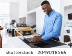 Positive black car salesman using laptop computer at work desk in auto dealership. Cheerful African American automobile manager working with pc in showroom store, free space