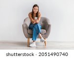 Happy Caucasian woman using cellphone, chatting on internet, working or studying online, sitting in armchair against white studio wall, full length. Cheery young lady watching video on mobile phone