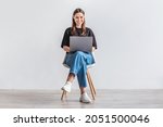 Happy young woman working online, sitting on chair and using laptop against white studio wall, full length. Positive Caucasian lady surfing internet, watching webinar on portable computer