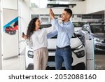 Emotional middle-eastern lovers young man and woman giving high five, standing by nice brand new car, holding key and laughing, buying new auto together at luxury showroom, copy space