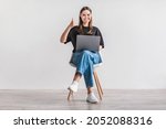 Smiling millennial woman working on laptop, showing thumb up gesture, recommending new website, sitting on chair against white wall. Young lady surfing internet, searching for information online
