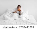 Small photo of Sad hungry caucasian young woman sits on white bed at home and eating burger, suffering from depression and mental problems, empty space. Night hunger, junk food, gluttony, substitution and stress