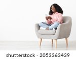 Cheerful african american young lady sitting in arm chair at home, using nice mobile application on brand new smartphone, shopping online, white background, panorama with copy space