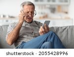 Poor Eyesight. Senior Man Squinting Eyes Reading Message On Phone Wearing Eyeglasses Having Problems With Vision Sitting On Couch At Home. Ophtalmic Issue, Bad Sight In Older Age Concept