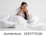 Small photo of Upset hungry european millennial woman eating big burger, sits on white bed in bedroom interior, has nutritional problems, obesity, food addiction. Unhealthy diet, bad eating habits, gluttony concept