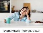 Sleepy young woman drinking coffee, feeling tired, suffering from insomnia and sleeping disorder. Sad female sitting in modern kitchen interior, empty space
