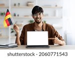 Small photo of Smiling arab guy student sitting at table athome, showing flag of Germany and laptop with blank screen, mockup, copy space. Positive middle-eastern young man studying German on Internet