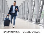 Online Check In. Handsome Arab Man Walking With Suitcase In Airport And Using Smartphone, Smiling Young Middle Eastern Guy Browsing Internet On Cellphone While Going To Flight Gate, Copy Space