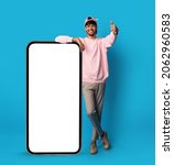 Small photo of Positive stylish indian guy posing with big smartphone with empty screen, showing thumb up and smiling, recommending newest mobile app, blue studio background, mockup, full length shot