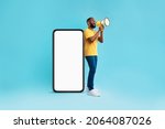 Happy young black guy shouting into megaphone, standing near big smartphone with empty screen, promoting your mobile app or website, offering mockup space for advertisement, blue background