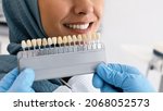 Small photo of Unrecognizable Stomatologist In Blue Gloves Holding Dental Teeth Shade Guide Chart, Choosing Right Enamel Color For Muslim Female Patient In Hijab During Stomatologic Treatment In Clinic, Closeup