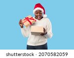 Christmas Gift. Joyful Oversized Black Lady Opening Xmas Present Box Standing Wearing Red Santa Hat Over Blue Background In Studio. New Year And Winter Holiday Celebration, Gifts Delivery