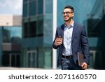 Cheerful middle eastern young businessman in glasses with laptop and coffee to go walking by street towards modern office building, going to job in the morning, panorama with copy space